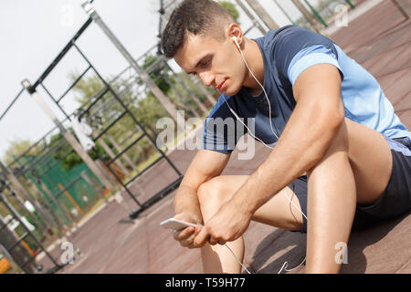 Gesunde Lebensweise. Junger Mann in Ohrhörer sitzen im freien Wahl Song auf dem Smartphone in der Nähe konzentrierten Stockfoto