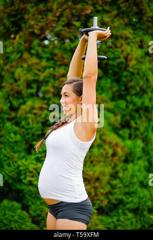 Schwangere Frau trainieren Trizeps Erweiterung mit Hantel in Park Stockfoto