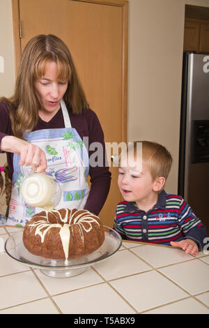 Mutter gießen weiße Schokolade auf Schokolade bundt Kuchen Glasur während drei Jahre alten Sohn Uhren. (MR) Stockfoto