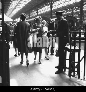 1960er Jahre, historisch, in einem Bahnhof, ein uniformierter Bahnsteig Wache oder Ticket Inspektor sammeln eine abfahrenden weiblichen Zug Fahrschein am Ausgang Gate, London Paddington. Stockfoto