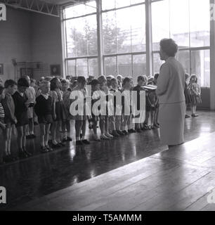 1960, historische, Schulleiterin Lesung einer Passage aus einem Buch - die Bibel - zu einer Gruppe der Grundschüler stehen in einer Linie am Morgen Montage, England, UK. Stockfoto