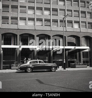 1964, historisch, ein Rolls-Royce-Auto mit Chauffeur, der vor dem Portland House, einem großen Bürogebäude in Victoria, London, geparkt ist. Der imposante 29-stöckige Turm wurde 1963 erbaut und war im Design „brutalistisch“, was der moderne, moderne Architekturstil der Zeit war. Stockfoto