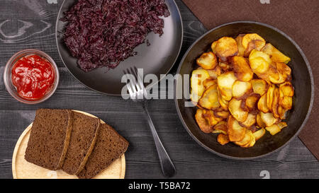 Fried Potato Chips in einem Topf mit dunkle Roggenbrot auf einer Holzplatte und hausgemachte gedünstetem Kohl, auf einem schwarzen Hintergrund im Landhausstil. Ansicht von oben Stockfoto