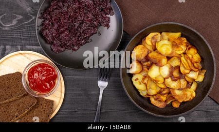 Fried Potato Chips in einem Topf mit dunkle Roggenbrot auf einer Holzplatte und hausgemachte gedünstetem Kohl, auf einem schwarzen Hintergrund im Landhausstil. Ansicht von oben Stockfoto