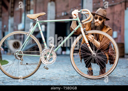 Bärtige hipster stilvoll gekleidet mit Hut stehend mit retro Fahrrad draußen auf der städtischen Hintergrund Stockfoto