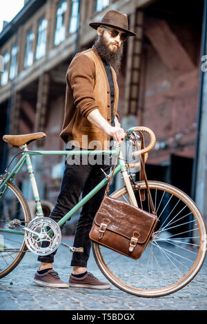 Bärtige hipster stilvoll gekleidet mit Hut stehend mit retro Fahrrad und Tasche im Freien auf der städtischen Hintergrund Stockfoto