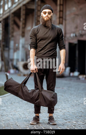 Porträt einer ernsten bärtiger Mann als Killer in Schwarz feste Kleidung, Tasche mit Waffen im Freien auf den industriellen Hintergrund gekleidet Stockfoto