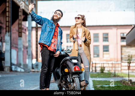 Stilvolle junger Mann und Frau, die selfie Foto beim zusammen stehen in der Nähe der retro Moped draußen auf der industriellen städtischen Hintergrund Stockfoto