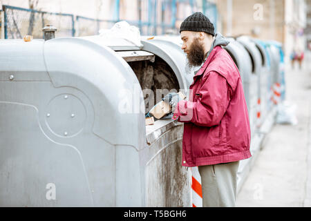 Obdachlose bärtige Bettler suchen einige Lebensmittel, die in den Papierkorb in der Stadt stöbern. Konzept der Armut und Arbeitslosigkeit Stockfoto