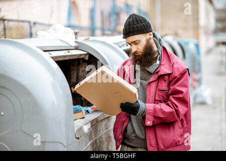 Obdachlose bärtige Bettler suchen einige Lebensmittel, die in den Papierkorb in der Stadt stöbern. Konzept der Armut und Arbeitslosigkeit Stockfoto