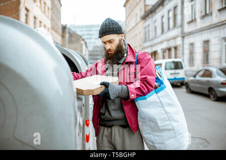 Obdachlose bärtige Bettler suchen einige Lebensmittel, die in den Papierkorb in der Stadt stöbern. Konzept der Armut und Arbeitslosigkeit Stockfoto