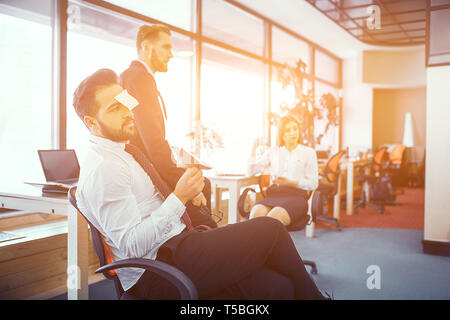 Bärtige Brünette mit Aufkleber auf seiner Stirn Leerlauf im sonnigen Büro. Holding Papier Arplane. Gelangweilt und Kollegen in der Nähe. Stockfoto