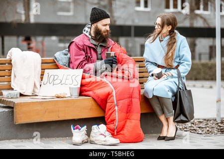 Obdachlose Bettler mit jungen Frau hören auf seine traurige Geschichte beim zusammen auf der Bank sitzen im Freien. Konzept eines menschlichen Verständnis Stockfoto