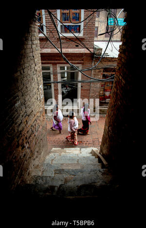 BHAKTAPUR, Nepal - April 5, 2019: Drei Frauen in traditioneller Kleidung Wandern in einer schmalen Straße, von einem dunklen Halle gesehen Stockfoto
