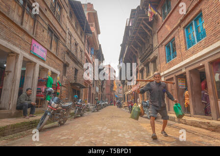 BHAKTAPUR, Nepal - April 5, 2019: Alte Mann Portierung Kunststoff Dosen wandern in einer engen Straße während eines Teils sonnig Nachmittag Stockfoto