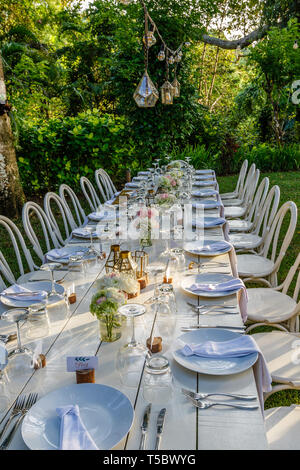 Set Table für eine Hochzeit Abendessen mit Rose Blumensträuße und Glas Lampen eingerichtet. Weiße Stühle aus Holz. Kalligraphie Namen tags. Tropische Hochzeit. Stockfoto