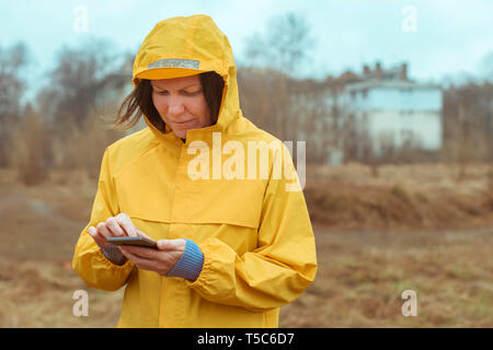 Frau im gelben Regenmantel SMS auf Handy im Freien an regnerischen Tag Stockfoto