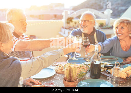 Gerne ältere Freunde Spaß jubeln mit Rotwein am Grill auf der Terrasse im Freien - Ältere Menschen das Abendessen toasten Gläser Stockfoto