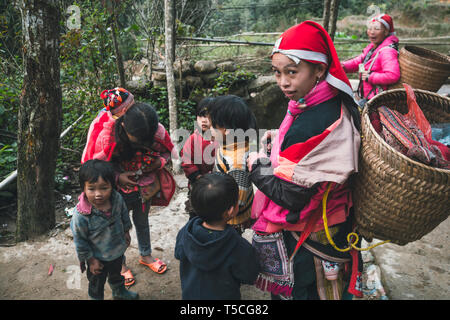 Mädchen aus dem Stamm der Roten Dzao, mit einem großen Weidenkorb hinter, von Kindern umgeben. TA PHIN, Lao Cai, VIETNAM - 12. Januar 2019 Stockfoto