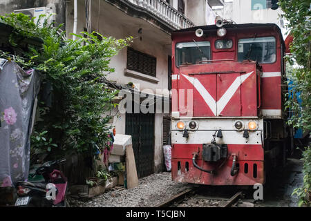 Ein roter Zug Lokomotive fährt obwohl eine schmale Straße in einem armen neighborhoood von Hanoi. Schmale Lücke zwischen Häuser ohne Sicherheitsmaßnahmen. Stockfoto