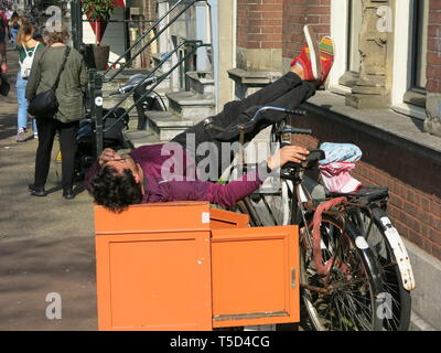 Eine Jugend liegt über einem strassenrand Kabinett mit seinen Füßen aufgestützt auf einer Fensterbank, während auf seinem Handy, das Leben auf der Straße in Amsterdam Stockfoto