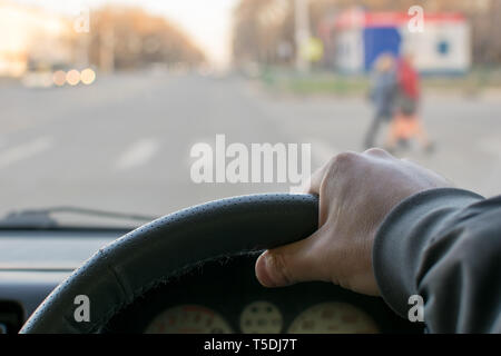 Blick aus dem Auto, Hand des Mannes am Lenkrad des Autos, befindet sich gegenüber der Fußgängerampel und Fußgänger über die Straße Stockfoto