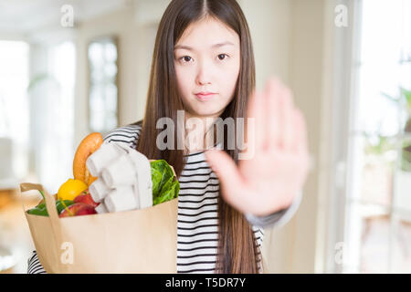 Schöne asiatische Frau mit papiertüte von frischen Lebensmitteln mit offener Hand das STOP-Schild mit ernsten und selbstbewussten Ausdruck, Verteidigung Geste Stockfoto