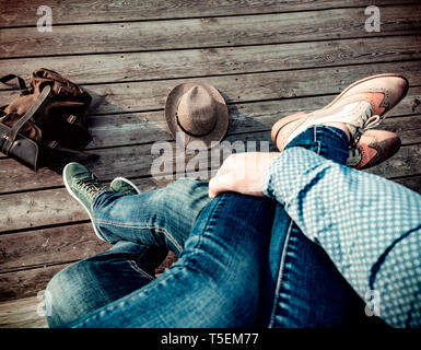 Paar sitzt Ihre Beine Umarmung auf Holzboden in Turnschuhe und Stiefel und Jeans auf den Außenbereich. Auf dem Boden liegen einen Strohhut und ein Stockfoto
