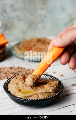 Nahaufnahme von einem Mann taucht ein Streifen von Karotte in einem hausgemachten Linse hummus gewürzt mit Paprika in einer grünen Keramik Teller serviert, auf einem weißen Holzmöbeln im Landhausstil Stockfoto