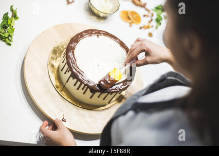 Der Konditor schmückt flüssige Schokolade Keks Kuchen mit weiße Creme, stehend auf einem Holzständer. Das Konzept der hausgemachte Kuchen, Kochen Kuchen. Stockfoto