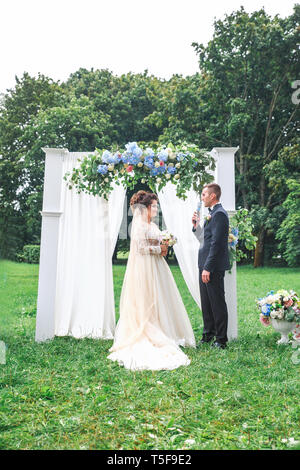 Der Bräutigam liest seinen Eid auf die Braut bei einer Zeremonie in der Nähe der Hochzeit Bogen unter freiem Himmel im Sommer. Stockfoto