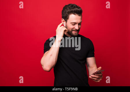 Portrait von Happy bearded hipster Mann Singen und Hören von Musik mit Handy und drahtlose Kopfhörer über rote Wand isoliert Stockfoto