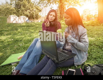 Paar der multi-ethnischen Mädchen Schüler sitzen auf Wiese Essen eines Apfels Stockfoto