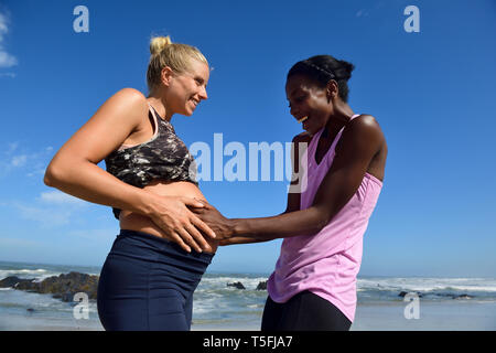 Glückliche Frau berühren Bauch der schwangeren Freundin am Strand Stockfoto