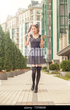Attraktive junge Frau mit Süßigkeiten in schwarzem Kleid zu Fuß auf der Straße Stockfoto