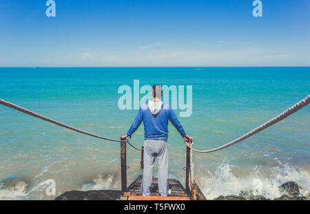 Kaukasische Mann steht auf alte hölzerne Treppe hinunter zum Mittelmeer im Morgen, Hintergrund Horizont, blaues Meer und Wellen Stockfoto