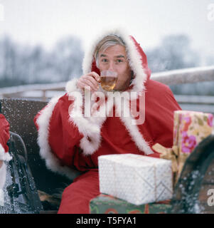 Joachim Fuchsberger trinkt einen Tee in einem Schlitten in einem Weihnachtsmann Kostüm, Deutschland 1970er. Joachim Fuchsberger Getränke Tee auf einem Schlitten in einem Santa Claus Kostüm, Deutschland 1970 s Stockfoto