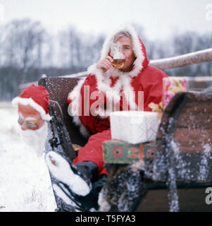 Joachim Fuchsberger trinkt einen Tee in einem Schlitten in einem Weihnachtsmann Kostüm, Deutschland 1970er. Joachim Fuchsberger Getränke Tee auf einem Schlitten in einem Santa Claus Kostüm, Deutschland 1970 s Stockfoto