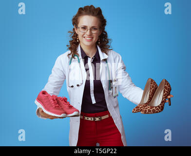 Lächelnd elegante Arzt Frau in bue Shirt, rote Hosen und weißen medizinische Gewand zeigen fitness Sneaker und High Heel Schuhe isoliert auf Blau. Stockfoto