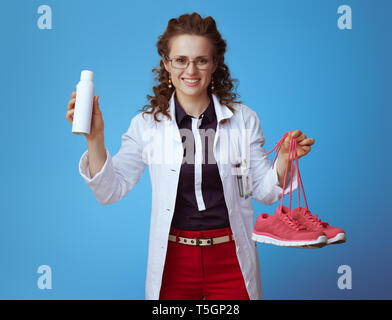 Happy Elegant Arzt Frau in bue Shirt, rote Hosen und weißen medizinische Gewand zeigen fitness Sneaker und Schuhe deodorizer Spray isoliert auf Blau backgrou Stockfoto