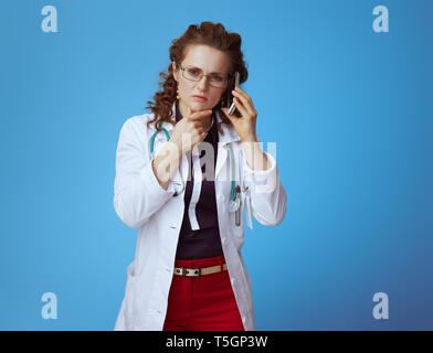 Nachdenklich elegante Arzt Frau in bue Shirt, rote Hosen und weißen medizinische Gewand sprechen auf einem Mobiltelefon auf blauem Hintergrund isoliert. Stockfoto