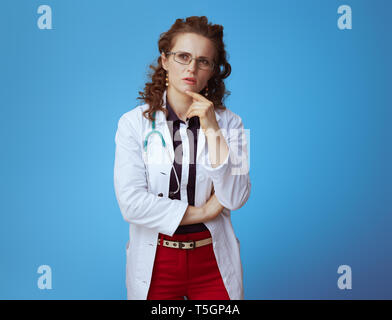 Portrait von nachdenklich moderne Arzt Frau in bue Shirt, rote Hosen und weißen medizinische Robe vor blauem Hintergrund. Stockfoto