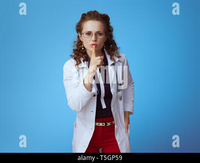 Portrait von eleganten Arzt Frau in bue Shirt, rote Hosen und weißen medizinische Gewand zeigen shh Geste gegen den blauen Hintergrund. Stockfoto