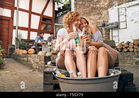 Freunde entspannen in einem Hinterhof in Sommer, junge Frauen kühlen ihre Füße in eine Wanne mit Getränken Stockfoto