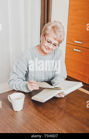 Ältere Frau liest Buch auf dem Tisch und trinken Kaffee, im Hause. Stockfoto