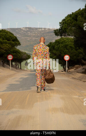 Rückansicht der glatzköpfige Mann mit Anzug mit bunten Polka-Dots zu Fuß auf der Landstraße mit Reisetasche Stockfoto