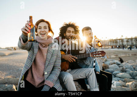 Drei glückliche Freunde mit Gitarre toasten Bierflaschen bei Sonnenuntergang Stockfoto