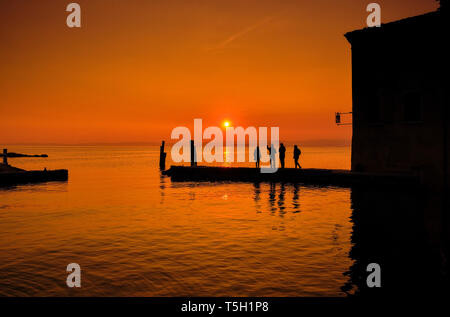 Italien, Punta San Vigilio, Silhouetten von vier Menschen auf der Jetty beobachten Sonnenuntergang über dem Gardasee im Winter Stockfoto