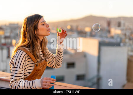 Junge Frau bläst Seifenblasen auf der Dachterrasse am Abend Stockfoto