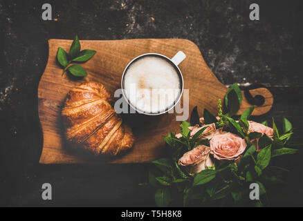 Tasse Cappuccino, Croissant und rosa Blüten auf schwarzem Hintergrund Stockfoto